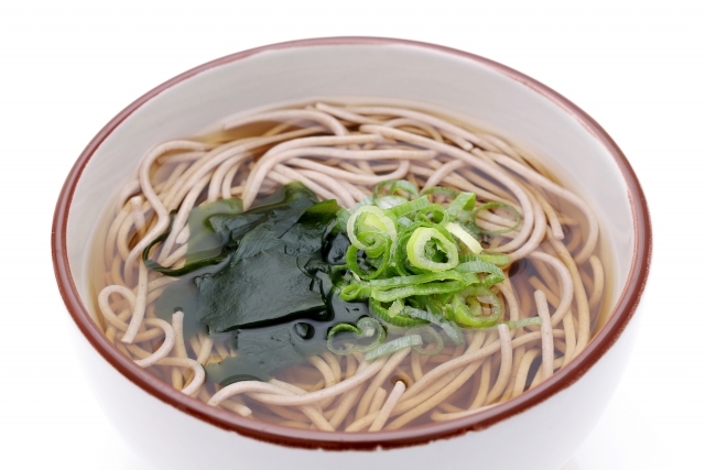 白い背景にフチが茶色のお椀に入った蕎麦の画像。トッピングはワカメと青ネギ。
